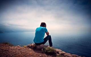 Man sitting rock ocean cloudy - liminal free wallpaper